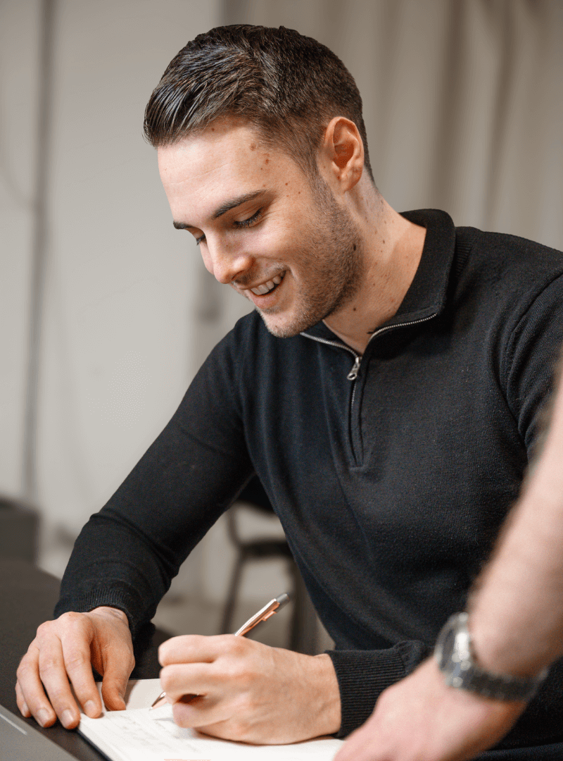 Ein junger Mann in einem schwarzen Pullover, der konzentriert etwas auf einem Papier schreibt.